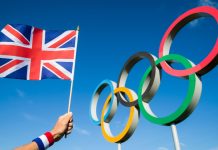 Team GB celebrates its roster with Topps partnership A hand of a Team GB supporter wearing red white and blue sweatband holds a UK flag in front of Olympic Rings.