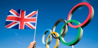 Team GB celebrates its roster with Topps partnership A hand of a Team GB supporter wearing red white and blue sweatband holds a UK flag in front of Olympic Rings.