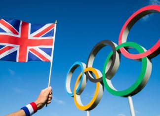 Team GB celebrates its roster with Topps partnership A hand of a Team GB supporter wearing red white and blue sweatband holds a UK flag in front of Olympic Rings.