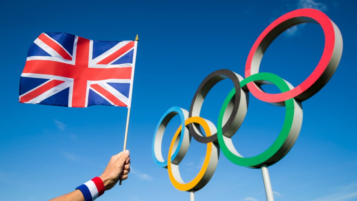 A hand of a Team GB supporter wearing red white and blue sweatband holds a UK flag in front of Olympic Rings.