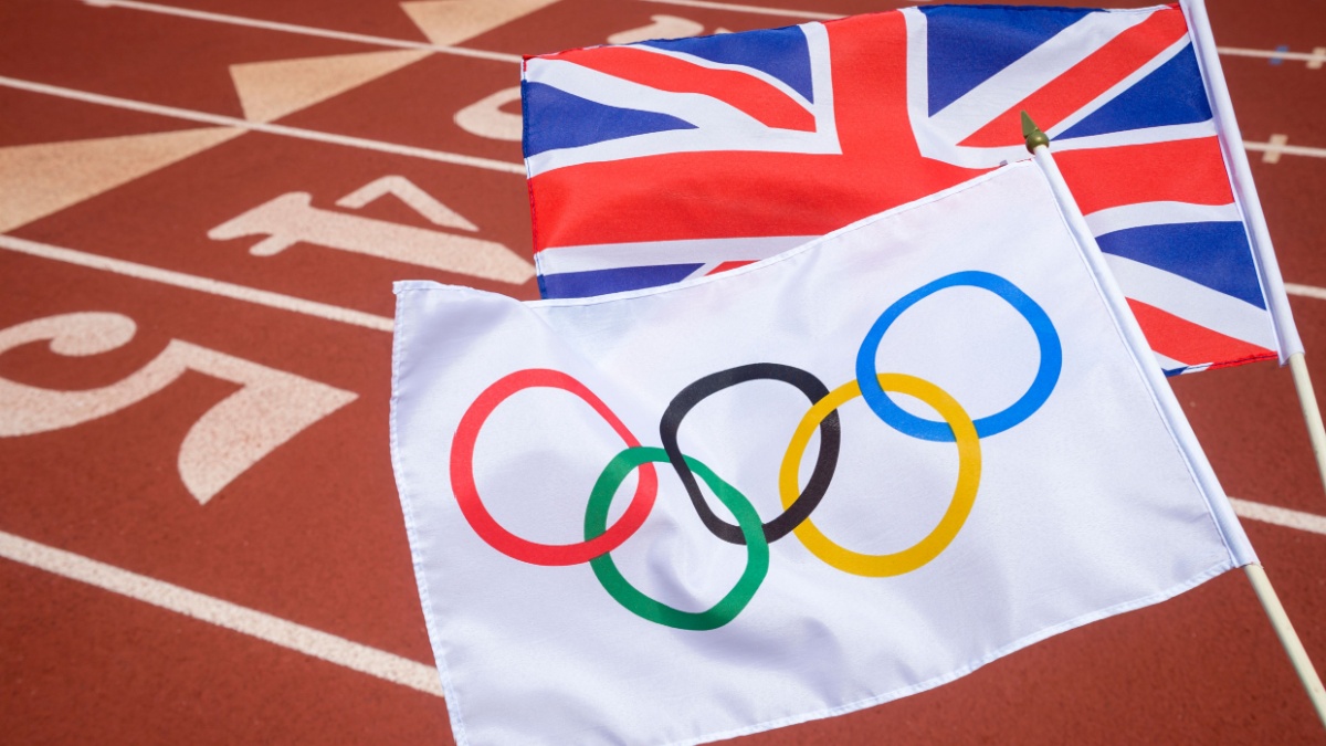 An Olympic and British union jack flag flutter together above the starting line of a red athletics track.