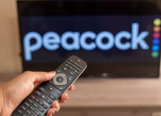 USL Super League chooses Peacock as official streaming platform A close-up of a hand holding a TV remote control seen in front of the Peacock logo.