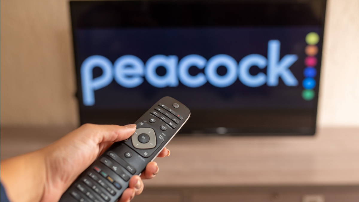 A close-up of a hand holding a TV remote control seen in front of the Peacock logo.