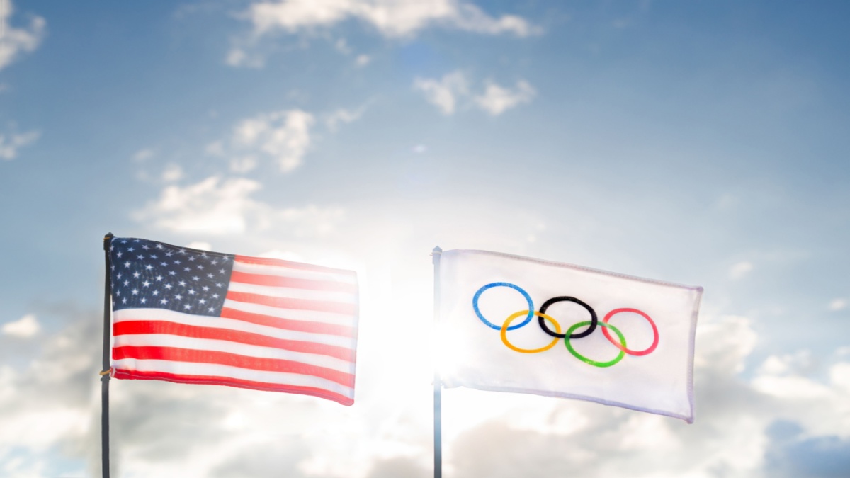 The iconic Olympic flag waves proudly beside the American flag under a vibrant sky.