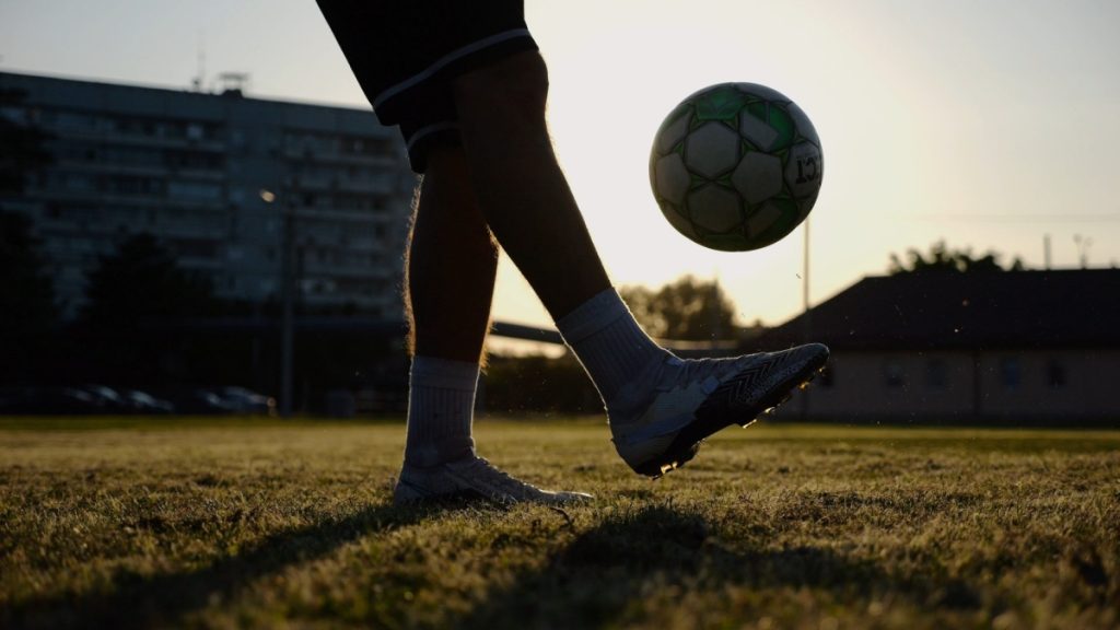 Legs of young man kicking fooball at green field. Male feet of professional footballer juggling soccer ball on stadium at sunset. Sportsman practicing tricks outdoor. Concept of a freestyle football.
