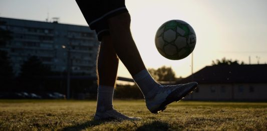 Legs of young man kicking ball at green field. Male feet of professional footballer juggling soccer ball on stadium at sunset. Sportsman practicing tricks outdoor. Concept of a freestyle football.