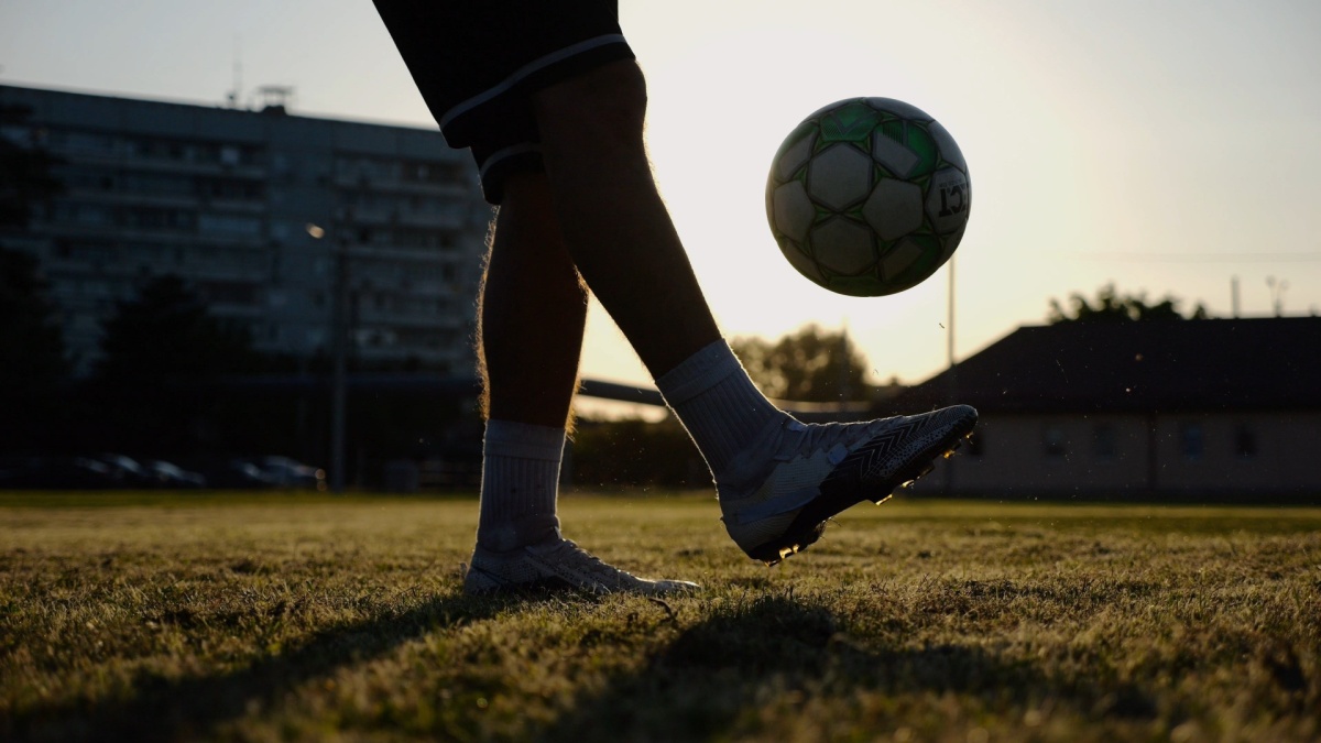 Legs of young man kicking fooball at green field. Male feet of professional footballer juggling soccer ball on stadium at sunset. Sportsman practicing tricks outdoor. Concept of a freestyle football.