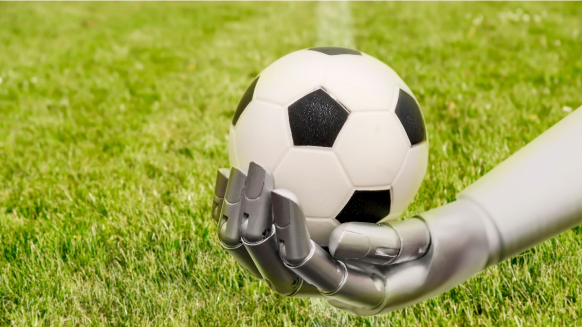 A robot arm reaches out to a football. Soccer playing with machine. The robot arm is in front of a soccer grass field.