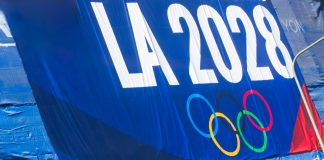 Who owns the future of the Olympic Games? A promotional banner for the LA 2028 Olympic Games featuring the Olympic rings, displayed on a blue background with a green fence below.