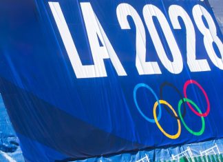 Who owns the future of the Olympic Games? A promotional banner for the LA 2028 Olympic Games featuring the Olympic rings, displayed on a blue background with a green fence below.