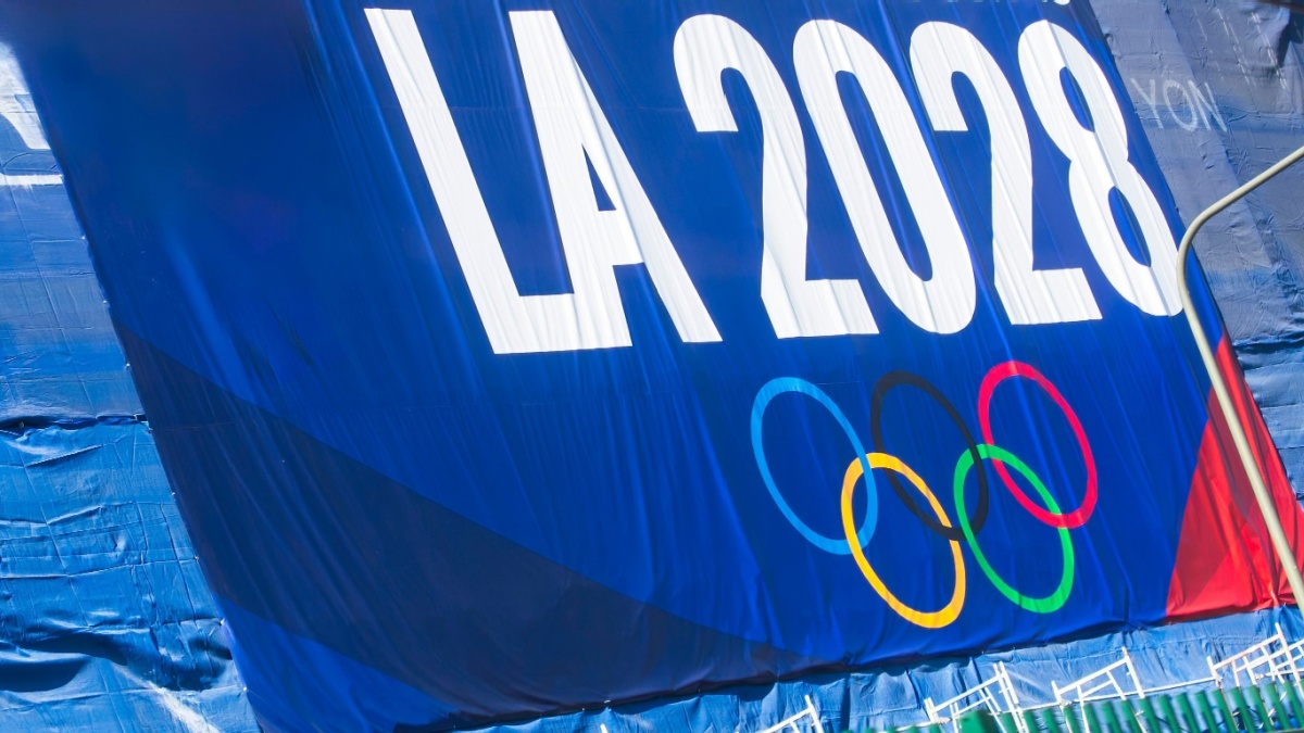 A promotional banner for the LA 2028 Olympic Games featuring the Olympic rings, displayed on a blue background with a green fence below.
