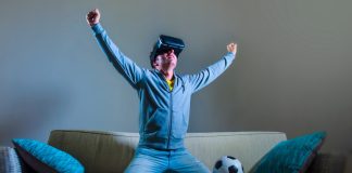 Burnley’s VR broadcast signals football’s next commercial goldmine young happy and excited gamer man using VR virtual reality goggles headgear set playing football simulation game celebrating scoring goal at home sofa couch enjoying 3D glasses fun experience.