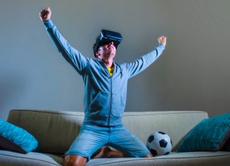 Burnley’s VR broadcast signals football’s next commercial goldmine young happy and excited gamer man using VR virtual reality goggles headgear set playing football simulation game celebrating scoring goal at home sofa couch enjoying 3D glasses fun experience.