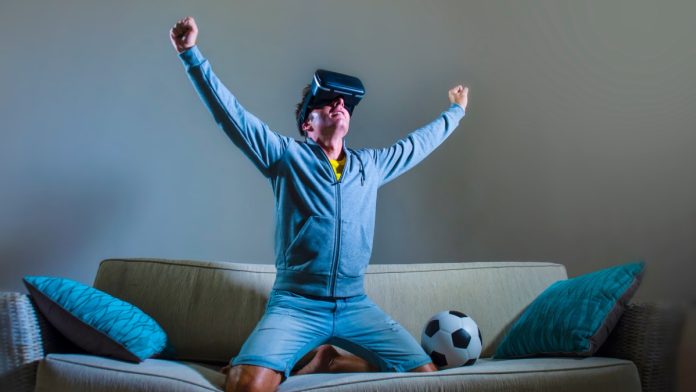young happy and excited gamer man using VR virtual reality goggles headgear set playing football simulation game celebrating scoring goal at home sofa couch enjoying 3D glasses fun experience.