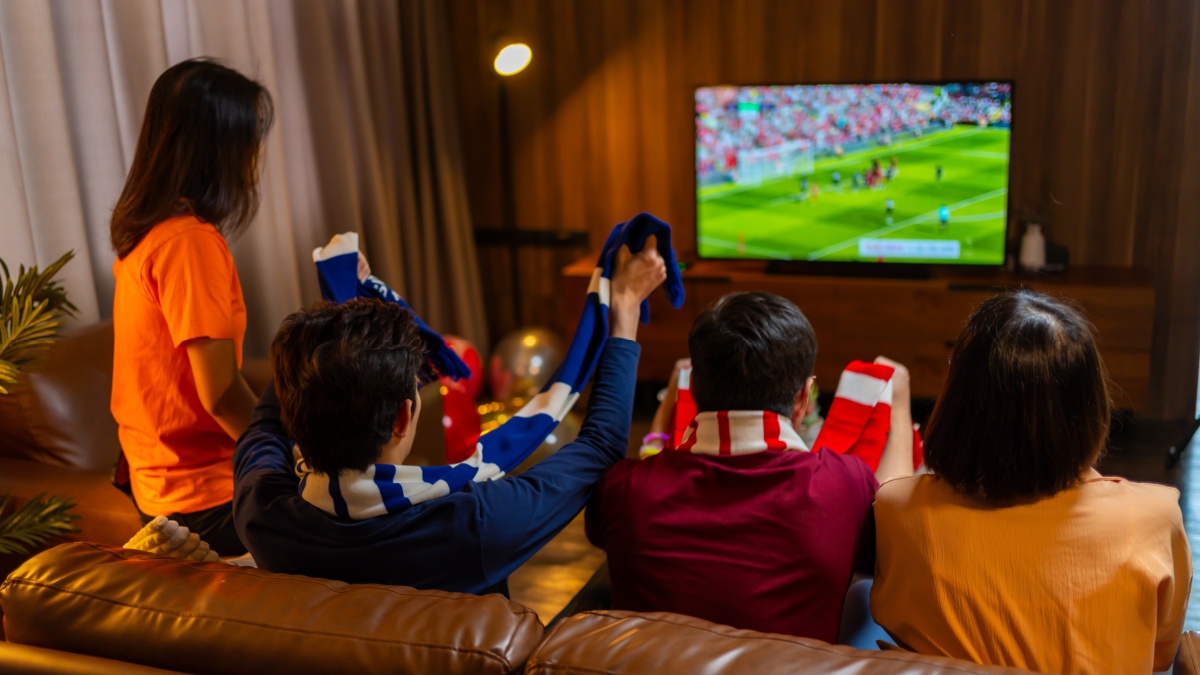 Group of Asian people friends sit on sofa watching and cheering football or soccer games competition on TV together at home.Happy man and woman sport fans celebrating sport team victory sports match.