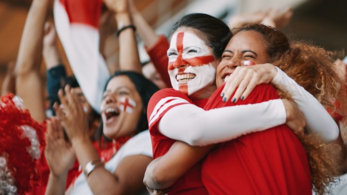 English female soccer fans with England flag painted on their faces hugging each other after their team's victory. English female spectators in football stadium celebrating their team's victory.