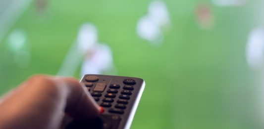 Man holding remote control for TV while there is football on television representing leisure watching sports.