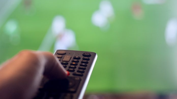 Man holding remote control for TV while there is football on television representing leisure watching sports.