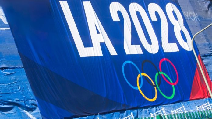 2024-11-20 Los Angeles USA A promotional banner for the LA 2028 Olympics featuring the Olympic rings, displayed on a blue background with a green fence below.