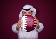 Could Qatar stage the inaugural Women’s Club World Cup? Arab man holding soccer ball in hand with Qatar flag standing on red background.