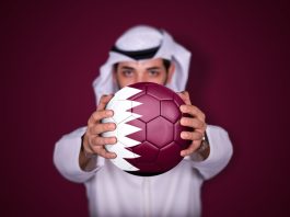 Could Qatar stage the inaugural Women’s Club World Cup? Arab man holding soccer ball in hand with Qatar flag standing on red background.