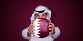 Could Qatar stage the inaugural Women’s Club World Cup? Arab man holding soccer ball in hand with Qatar flag standing on red background.
