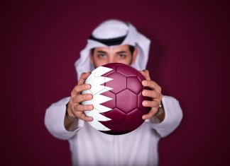 Could Qatar stage the inaugural Women’s Club World Cup? Arab man holding soccer ball in hand with Qatar flag standing on red background.