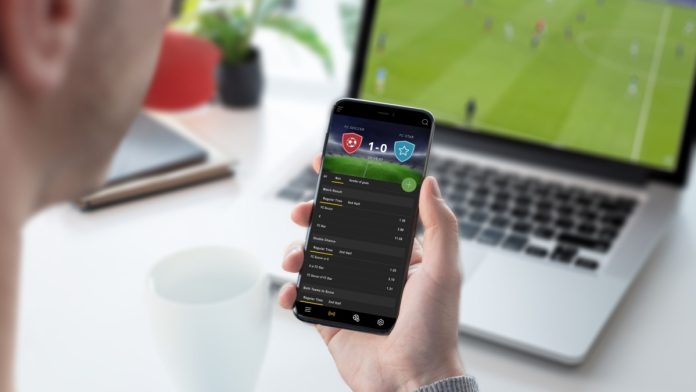 Guy focused on betting odds on his smartphone. Follows a football game on his laptop
