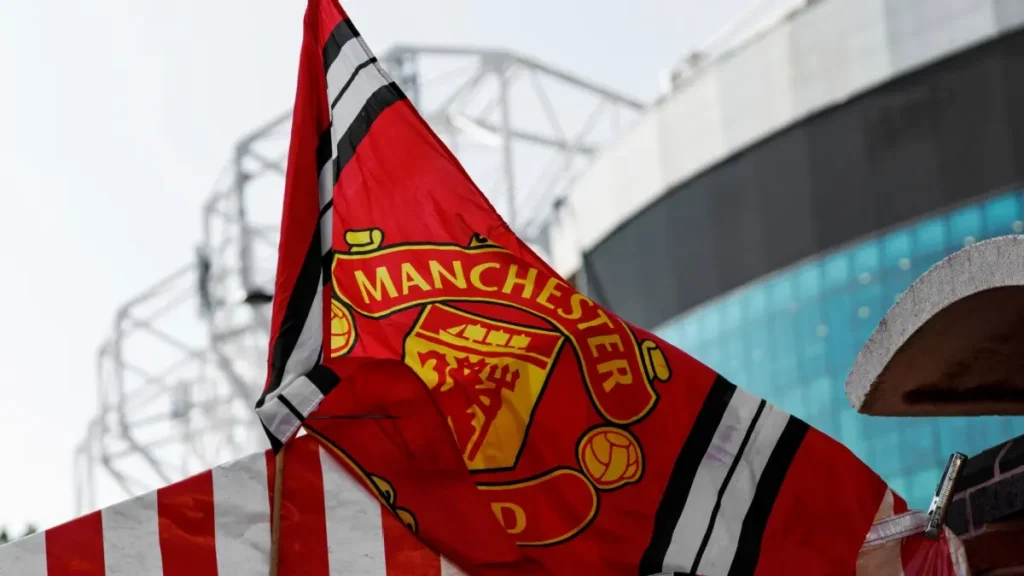 Red Manchester United flag with club crest blows in the wind near Old Trafford stadium.